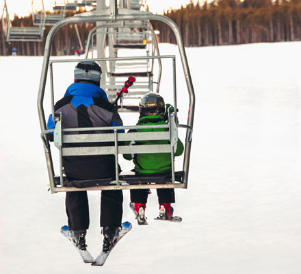 Small boy and his father sitting on a chair lift in late afternoon light in winter, viewed from behind