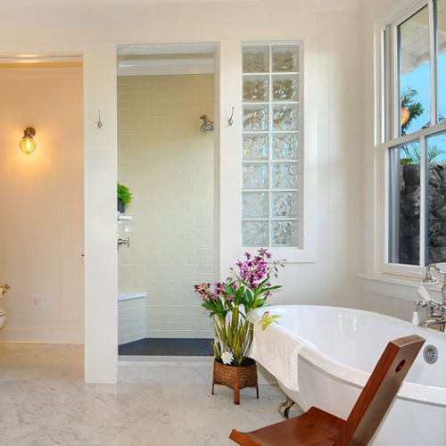 Clean, modern white bathroom with soaking tub, marble double vanities, walk-in shower and marble flooring. Large windows look out to handcrafted Hawaiian rock wall to outdoor shower. 