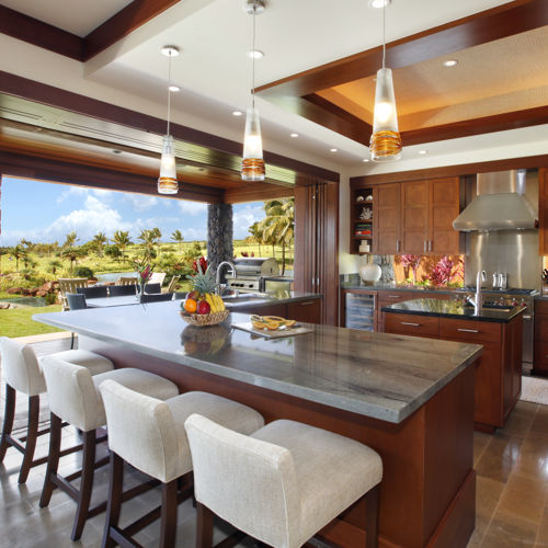 Modern open chef's kitchen with island and L-shaped counter lined with four bar stools. Entire wall slides open to covered lanai, plunge pool, and outdoor seating.