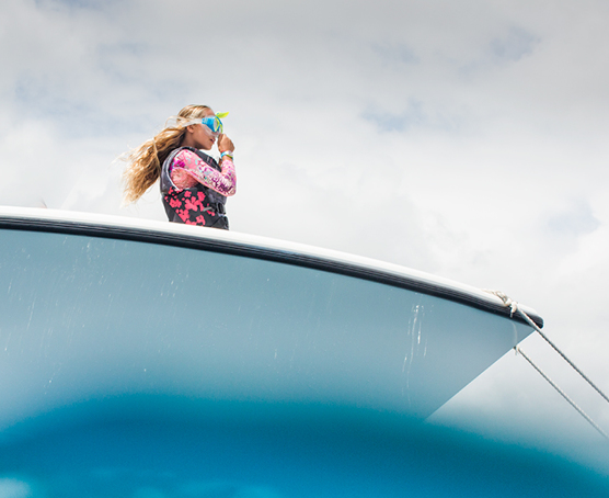 A girl in a long-sleeve rash guard and snorkel mask standing at the front of the boat. View from the water looking up at the boat. 