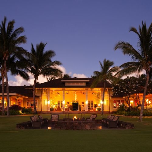 Panoramic view across lawn of warm glow of lights in the Kukui'ula Club House at dusk.